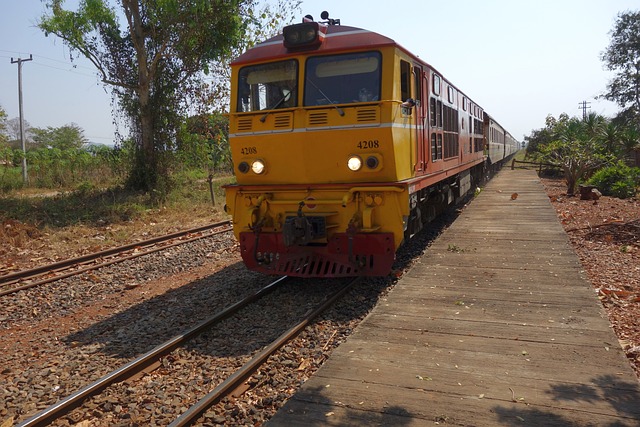 Un train jaune et rouge de la Thaïlande à l'arrêt sur les voies ferrées, avec une petite plateforme en bois et des arbres en arrière-plan, en zone rurale.