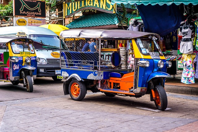 Deux tuk-tuks bleus et jaunes garés dans la rue animée de Khao San Road à Bangkok, avec des enseignes de bars et de magasins en arrière-plan.