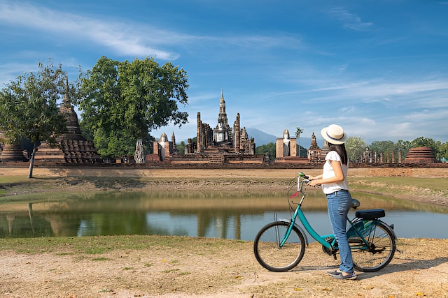 Une femme portant un chapeau se tient à côté de son vélo turquoise et admire la vue sur les ruines du temple Wat Mahathat de l'autre côté d'un lac, dans le parc historique de Sukhothai.