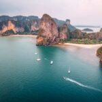 Vue aérienne des falaises karstiques de la plage de Railay à Krabi, en Thaïlande, avec des bateaux sur une mer turquoise et une plage de sable blanc.