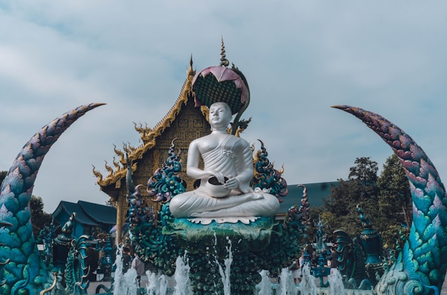 Statue blanche du Bouddha devant l'ubosot du Temple Bleu (Wat Rong Suea Ten) à Chiang Rai, avec ses sculptures de nagas bleus et dorés.