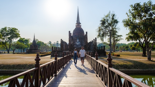 Un couple se tenant la main marche sur un pont en bois en direction des ruines du temple Wat Sa Si, avec son chedi de style sri-lankais, dans le parc historique de Sukhothai.
