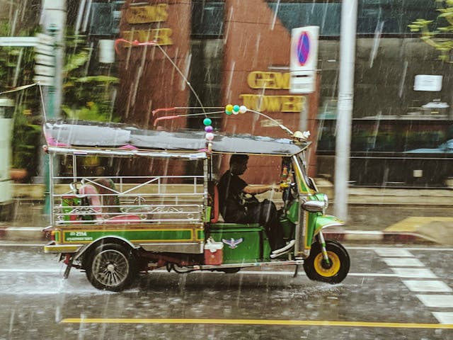 Tuk-tuk vert emblématique roulant sous la pluie en Thaïlande, illustrant les arnaques courantes à éviter par les touristes.