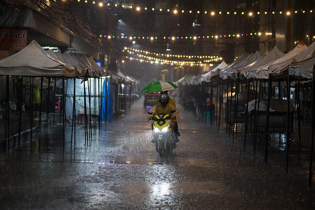 Scène de rue à Bangkok la nuit sous une pluie battante, avec un scooter et son passager circulant dans une allée de marché.