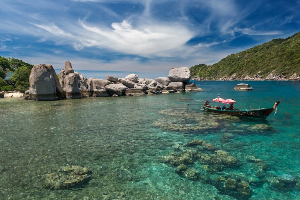 Bateau taxi traditionnel thaïlandais naviguant sur l'eau turquoise d'une baie de Koh Tao, avec des rochers de granit et des récifs coralliens visibles sous la surface.