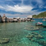 Bateau taxi traditionnel thaïlandais naviguant sur l'eau turquoise d'une baie de Koh Tao, avec des rochers de granit et des récifs coralliens visibles sous la surface.