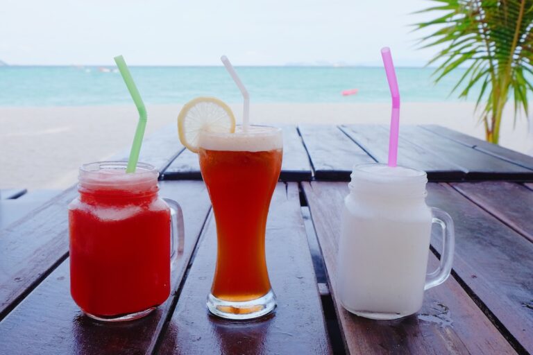 Trois verres de boissons thaïlandaises colorées, un jus de fruit, un thé glacé et un smoothie coco, sur une table face à la mer.