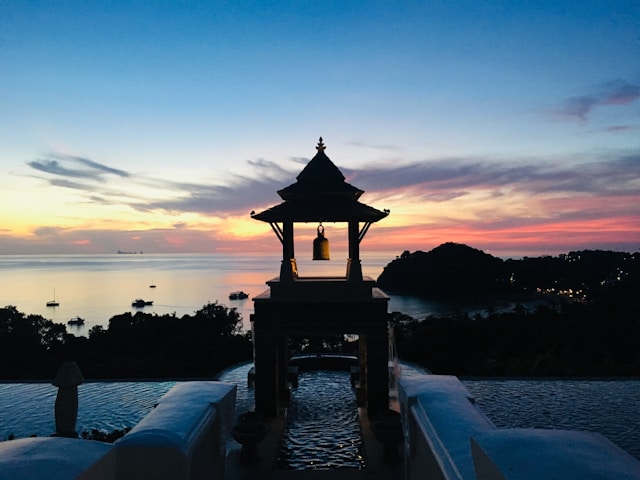 Vue sur la mer d'Andaman au crépuscule depuis une piscine à débordement à Koh Lanta, avec un pavillon thaïlandais et une cloche en silhouette.