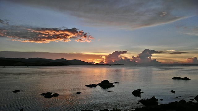 Magnifique coucher de soleil sur la mer en Thaïlande, avec des nuages colorés, des rochers au premier plan et des montagnes au loin.