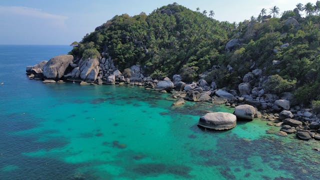 Vue aérienne d'une baie isolée de Koh Tao, montrant des eaux turquoise transparentes qui révèlent des récifs, bordée par des rochers de granit et une colline recouverte de jungle.