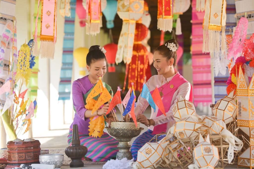 Deux femmes en costume traditionnel thaïlandais sourient en préparant des décorations pour un festival, illustrant la richesse de la culture et du savoir-vivre en Thaïlande.