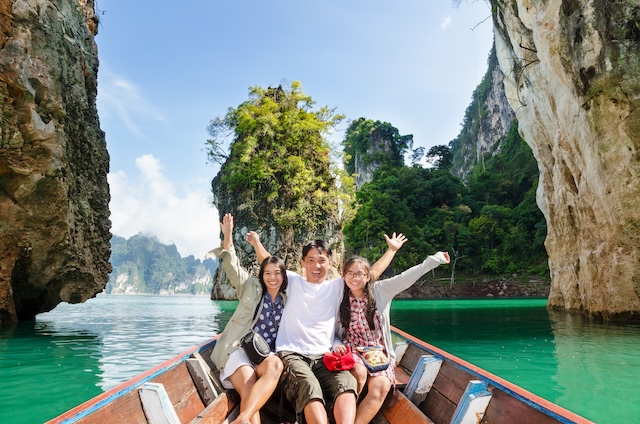 Famille souriante (père, mère et fille) assise à l'avant d'un bateau traditionnel en bois, levant les bras en signe de joie, naviguant sur une eau turquoise entourée de falaises karstiques verdoyantes en Thaïlande.