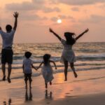 Silhouette d'une famille avec deux enfants sautant et courant joyeusement sur une plage au bord de la mer, avec le soleil se couchant sur l'horizon.
