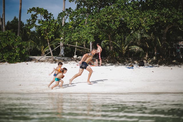 Un homme et deux jeunes enfants en maillots de bain courent et s'amusent sur le sable blanc au bord de l'eau, sur une plage tropicale en Thaïlande.