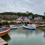 Vue d'ensemble du Fisherman Village à Pattaya, montrant des bateaux de pêche colorés sur un plan d'eau devant des bâtiments de style scandinave et des terrasses de restaurants.