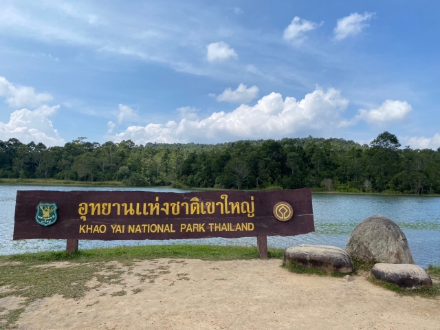 Panneau officiel en bois du parc national de Khao Yai devant un lac paisible, avec une forêt dense en arrière-plan sous un ciel bleu.