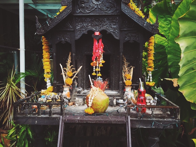 Maison des esprits traditionnelle en bois sombre, ornée de guirlandes de fleurs jaunes et d'offrandes quotidiennes comme une noix de coco, de l'encens et une boisson rouge, dans un jardin en Thaïlande.