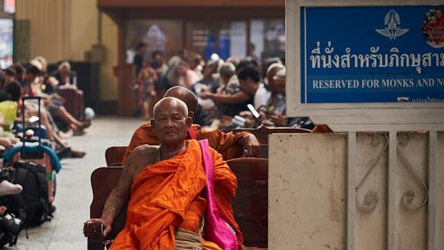 Un moine bouddhiste âgé avec des tatouages traditionnels, assis sur un banc en bois dans une gare thaïlandaise à côté d'un panneau lui réservant la place.