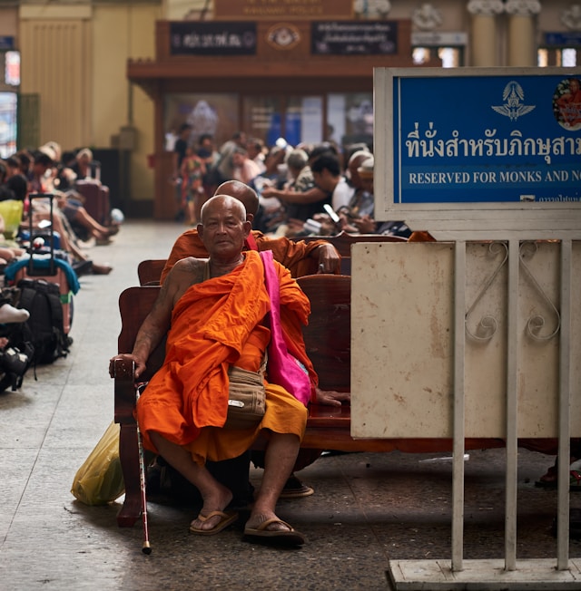 Un moine bouddhiste âgé avec des tatouages traditionnels, assis sur un banc en bois dans une gare thaïlandaise à côté d'un panneau lui réservant la place.