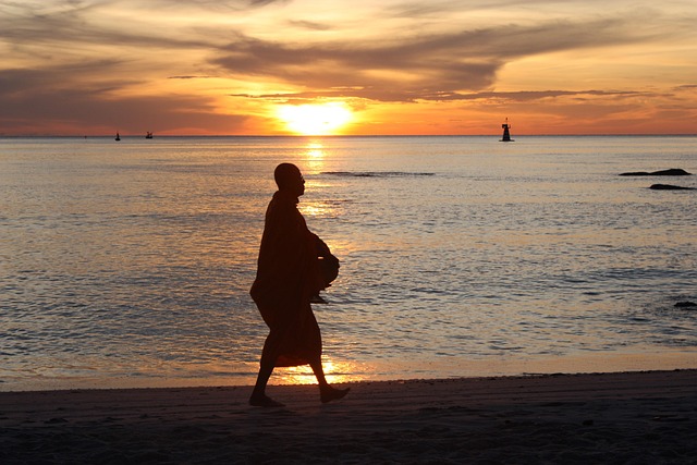 Silhouette d'un moine bouddhiste marchant sur une plage en Thaïlande au coucher du soleil, symbolisant la paix d'un voyage réussi.