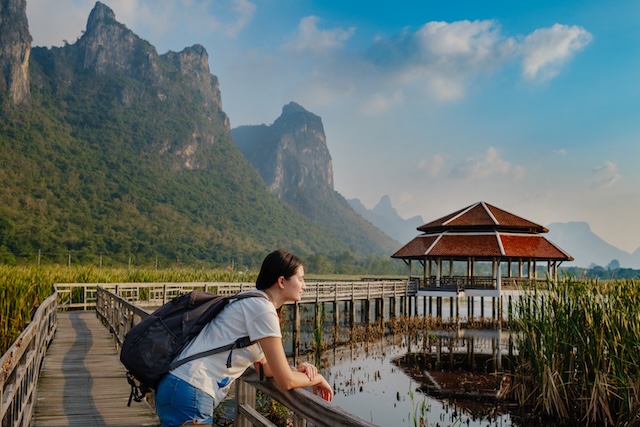 Une voyageuse avec un sac à dos, accoudée à une balustrade en bois, regardant un pavillon thaïlandais sur un lac marécageux, avec les montagnes karstiques du parc national de Khao Sam Roi Yot en arrière-plan.