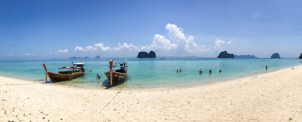 Vue panoramique d'une plage de sable blanc à Koh Lanta avec des bateaux traditionnels à longue queue sur une mer turquoise.