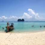 Vue panoramique d'une plage de sable blanc à Koh Lanta avec des bateaux traditionnels à longue queue sur une mer turquoise.