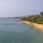 Vue panoramique d'une longue plage de sable en Thaïlande bordée de palmiers et d'une mer calme, image de couverture pour un guide sur le budget de voyage.