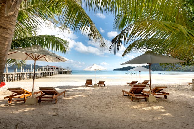 Vue d'une plage de sable blanc en Thaïlande avec des transats et parasols sous les palmiers, un ponton en bois et la mer calme en arrière-plan.