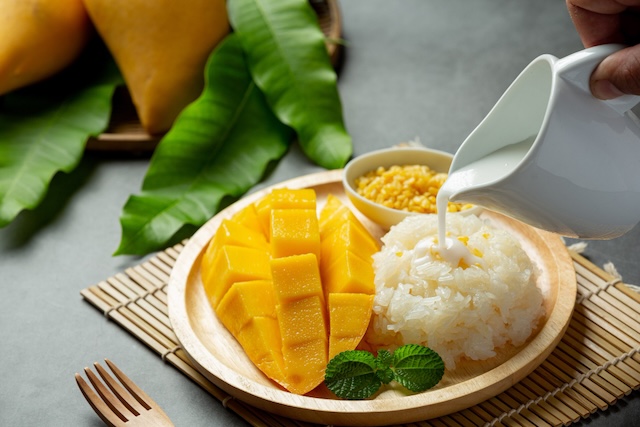 Assiette de riz gluant à la mangue fraîchement coupée, avec une main versant du lait de coco sur le riz, dessert typique de Thaïlande.