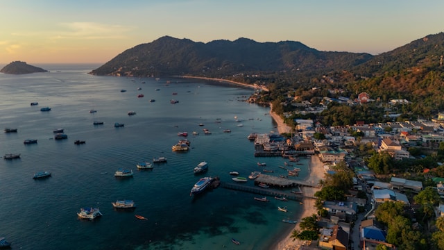 Vue aérienne de la baie de Sairee à Koh Tao au coucher du soleil, montrant de nombreux bateaux de plongée et bateaux-taxis ancrés dans l'eau, avec le village côtier et les collines verdoyantes en arrière-plan.