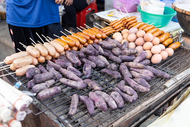 Stand de nourriture de rue en Thaïlande montrant des patates douces violettes, des saucisses et des œufs en train de griller sur un barbecue.