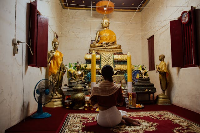 Personne vue de dos portant un pantalon blanc et un châle couvrant les épaules, agenouillée en prière devant des statues de Bouddha dans un temple thaïlandais.