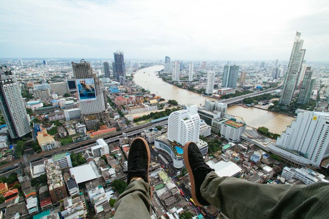 Vue à la première personne depuis le sommet d'un immeuble à Bangkok, avec les pieds surplombant la ville et le fleuve Chao Phraya, illustrant la vie d'un expatrié en Thaïlande.
