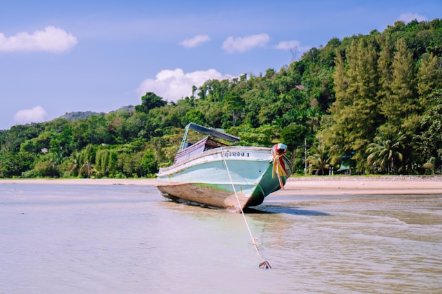 Un bateau de pêche thaïlandais de couleur verte posé sur le sable d'une plage à marée basse, avec une colline verdoyante en arrière-plan.