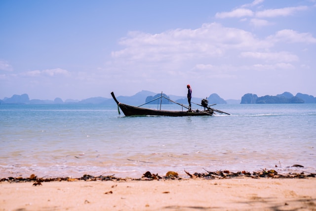 Un homme coiffé d'un bonnet rouge se tient sur son bateau long-tail en bois près d'une plage de sable, avec les îles de la baie de Phang Nga au loin.