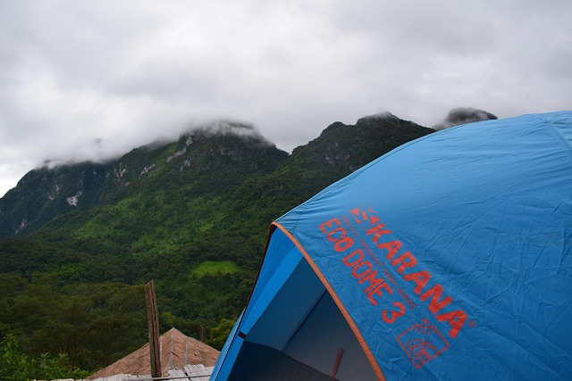 Tente bleue de camping au premier plan avec les montagnes verdoyantes et nuageuses de Doi Chiang Dao en arrière-plan.