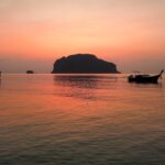 Vue de la baie de Phang Nga au coucher du soleil, avec la silhouette d'un karst calcaire et de bateaux long-tail sur une mer calme.