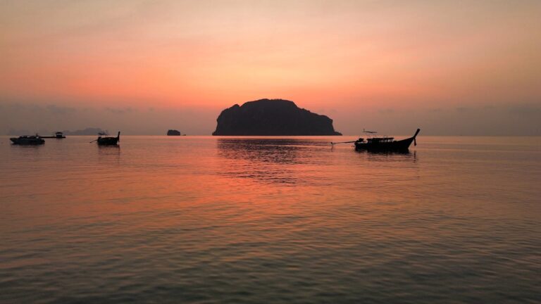 Vue de la baie de Phang Nga au coucher du soleil, avec la silhouette d'un karst calcaire et de bateaux long-tail sur une mer calme.