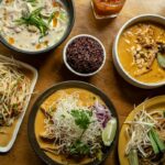 Table en bois vue de dessus avec un assortiment de plats thaïlandais : Som Tam, Khao Soi, Pad Thai et divers currys.