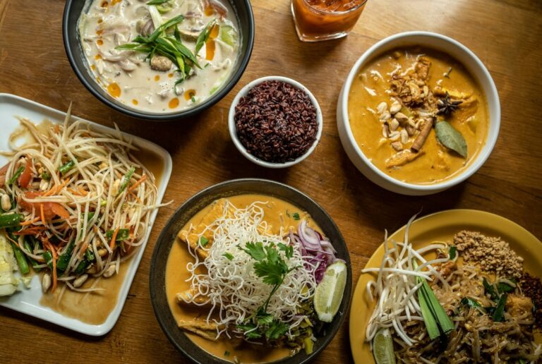 Table en bois vue de dessus avec un assortiment de plats thaïlandais : Som Tam, Khao Soi, Pad Thai et divers currys.