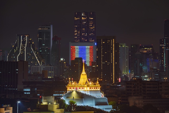 ue nocturne de Bangkok avec le drapeau thaïlandais illuminé sur un gratte-ciel et le temple Wat Saket (Montagne d'Or) au premier plan.