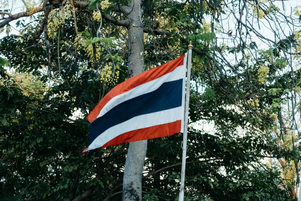 Le drapeau national de la Thaïlande, le Thong Trairong, flottant dans un jardin avec des arbres en arrière-plan.