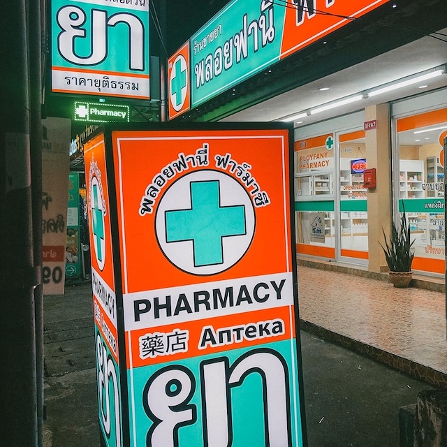 Enseigne lumineuse et colorée d'une pharmacie de rue en Thaïlande la nuit, avec des inscriptions en thaï, anglais, russe et japonais.