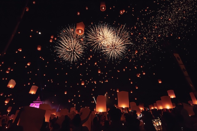 Des milliers de lanternes célestes s'élevant dans le ciel nocturne au-dessus d'une foule pendant le festival Yi Peng à Chiang Mai, avec des feux d'artifice en arrière-plan.