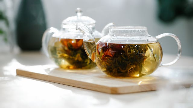 Deux théières en verre transparentes contenant une infusion chaude de feuilles et de fleurs de plantes médicinales thaïlandaises, posées sur un plateau en bois dans une atmosphère lumineuse et apaisante.