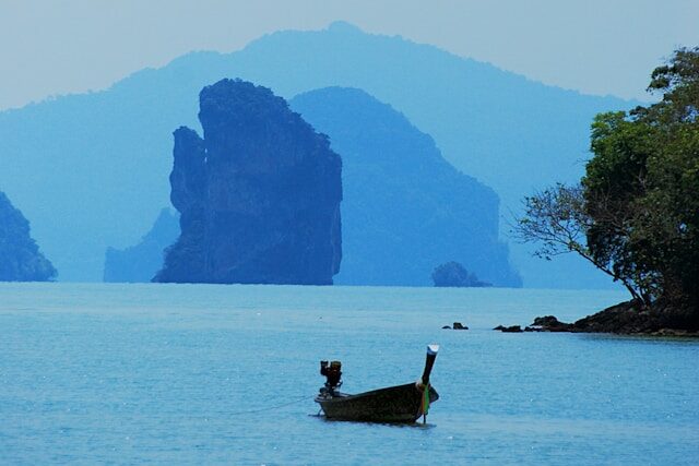 Un bateau long-tail thaïlandais traditionnel flottant sur une mer bleue, avec en arrière-plan les karsts calcaires de la baie de Phang Nga dans la brume.