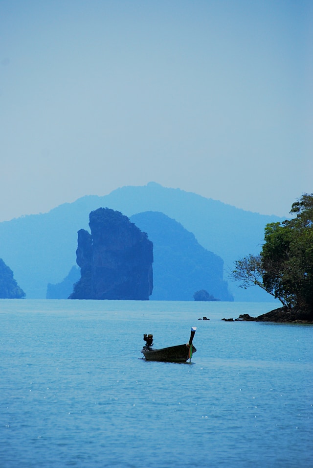 Un bateau long-tail thaïlandais traditionnel flottant sur une mer bleue, avec en arrière-plan les karsts calcaires de la baie de Phang Nga dans la brume.