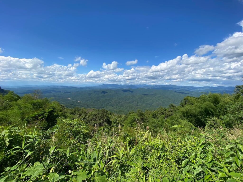 Panorama des montagnes verdoyantes de Nan en Thaïlande depuis un point de vue, avec une végétation luxuriante au premier plan sous un ciel bleu nuageux.