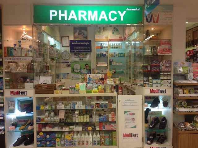 Devanture d'une pharmacie locale thaïlandaise bien achalandée, avec une enseigne verte 'PHARMACY' et des étagères remplies de médicaments et de produits de parapharmacie.